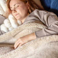 A woman laying underneath a tan etra-fluffy throw.