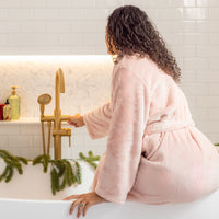 Woman in a pink robe standing by a bathtub with gold fixtures and greenery.