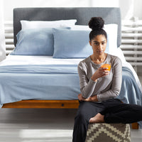 Woman sitting infront of a bed with a glass of orange juice in her hand and a polar blue Polartec Softec Microfleece blanket draped over the bed.