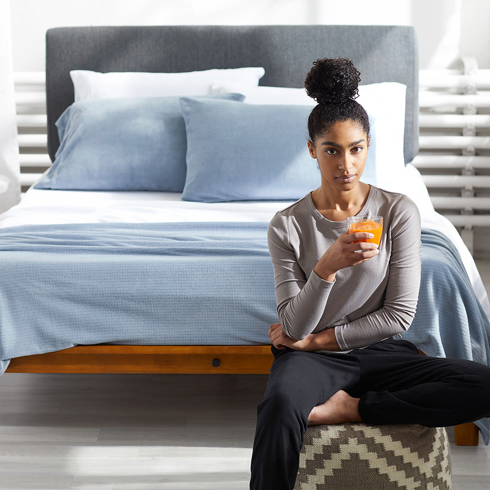 Woman sitting infront of a bed with a glass of orange juice in her hand and a polar blue Polartec Softec Microfleece blanket draped over the bed.