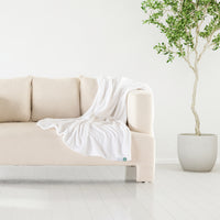 A white velvety plush throw draped over the arm of a beige sofa next to a plant.