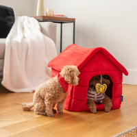 Two dogs sitting inside and near a red peanuts snoopy dog house pet bed.