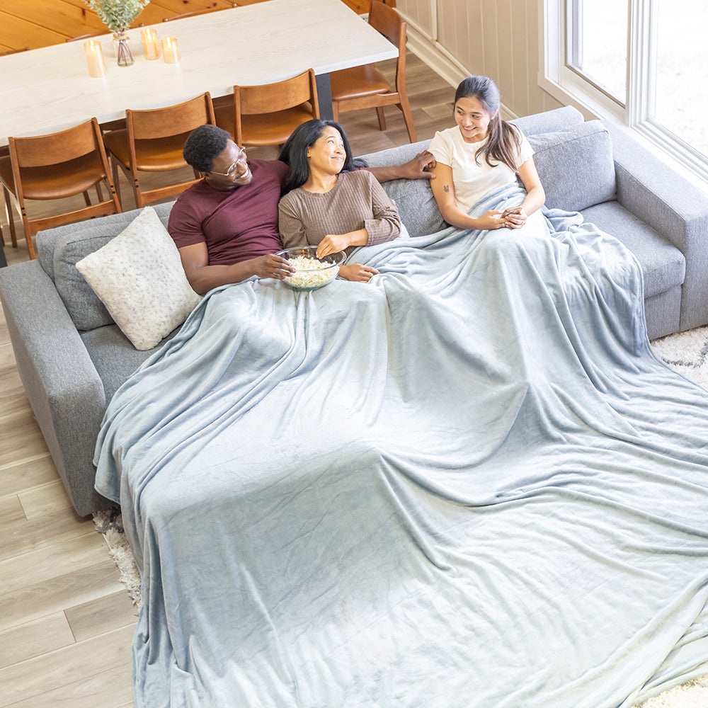 3 people sitting under a blue velvety plush family blanket on a grey sofa.