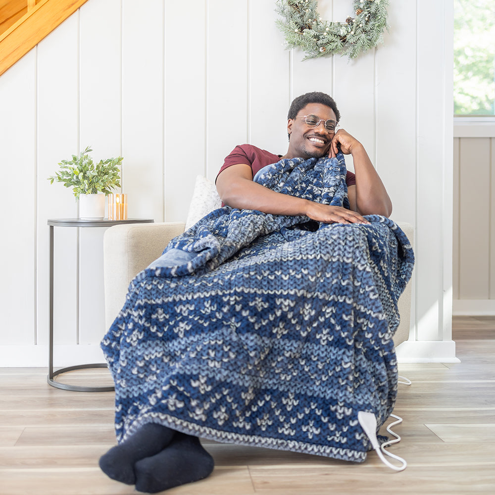 A man sitting on a chair, wrapping himself in a blue heated throw in a bright white room.
