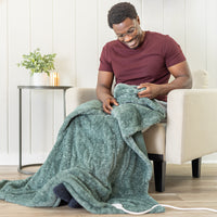 A man sitting on a chair wrapped in a soft, fluffy green heated throw in a bright white room.