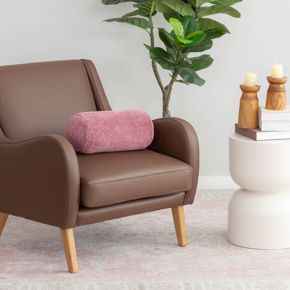 A pink EcoThread Neckroll Pillow sitting on a brown leather chair next to a plant and table.