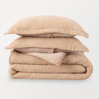A single retro plush comforter folded and stacked with two matching pillow shams stacked in a pile. Almond Seed color is a tan color.
