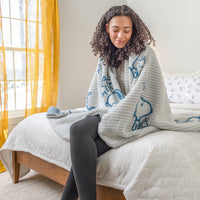 Woman sitting on a bed wrapped in a blanket with a blue pattern, in a room with yellow curtains.