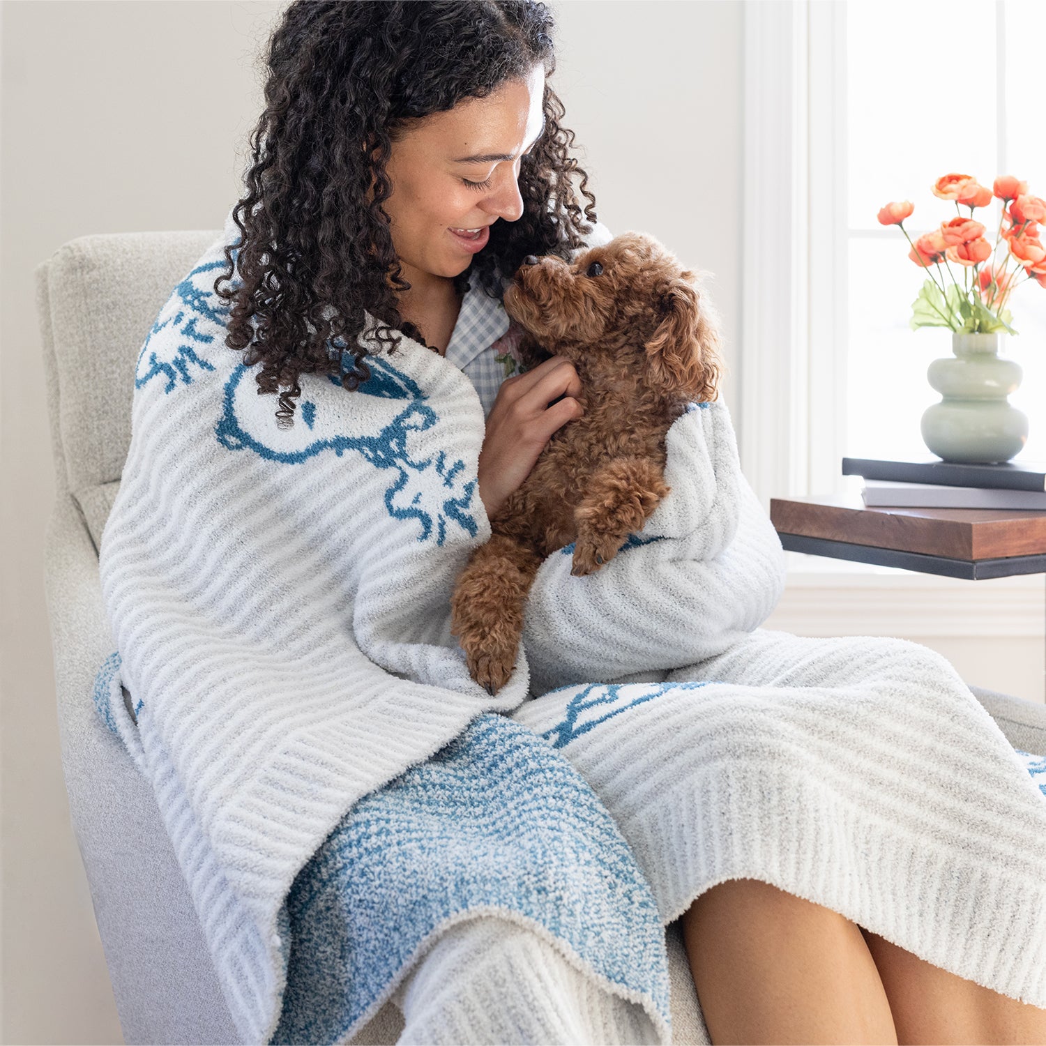 A woman sitting on a chair holding a dog with our Peanuts MellowSoft Throw wrapped around her shoulders.