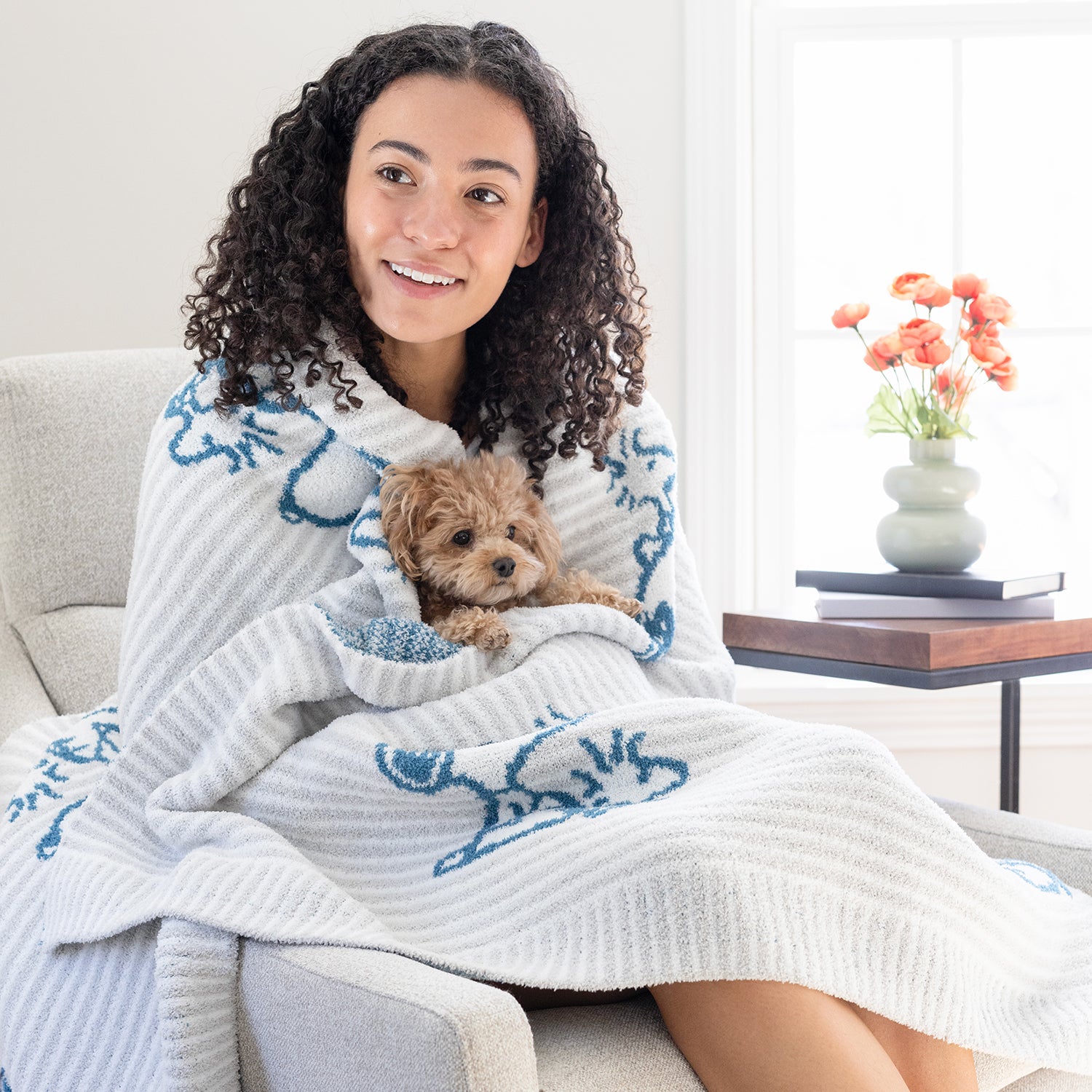 A woman sitting on a chair holding a dog with our Peanuts MellowSoft Throw wrapped around her shoulders.