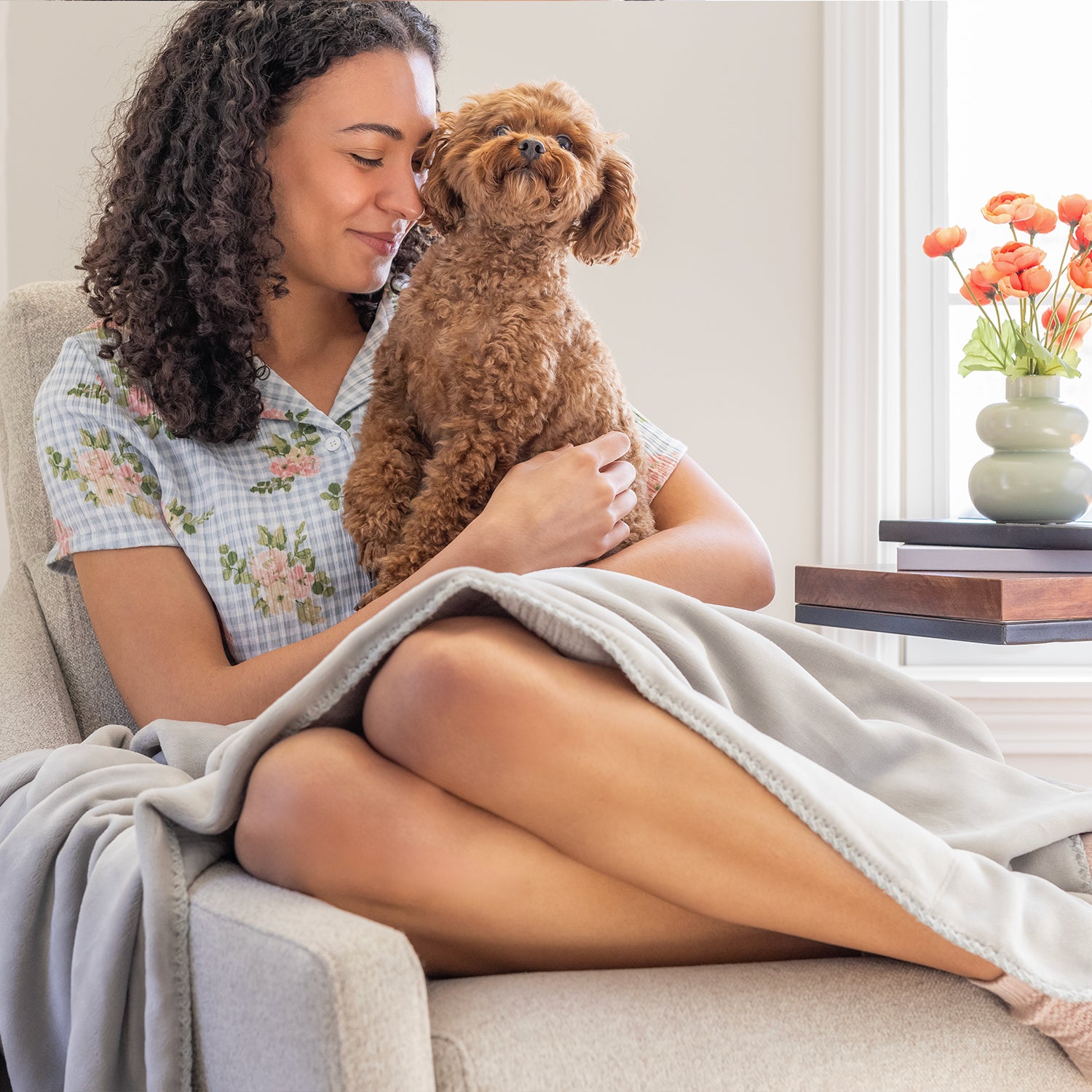 A woman sitting on a grey chair with a dog and a grey throw draped over her legs.
