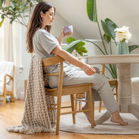 Woman sitting at a table holding a mug in a cozy room with plants and a vase.