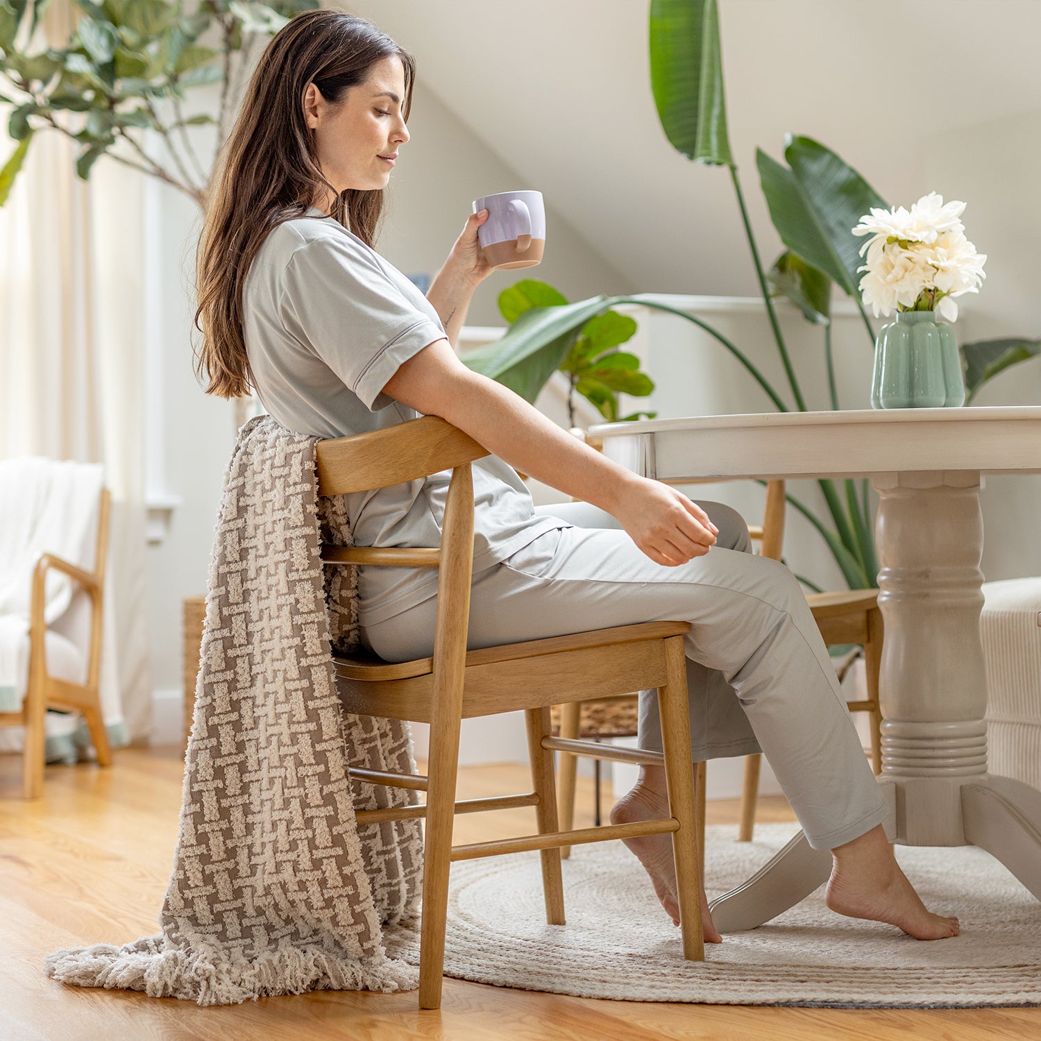 Woman sitting at a table holding a mug in a cozy room with plants and a vase.