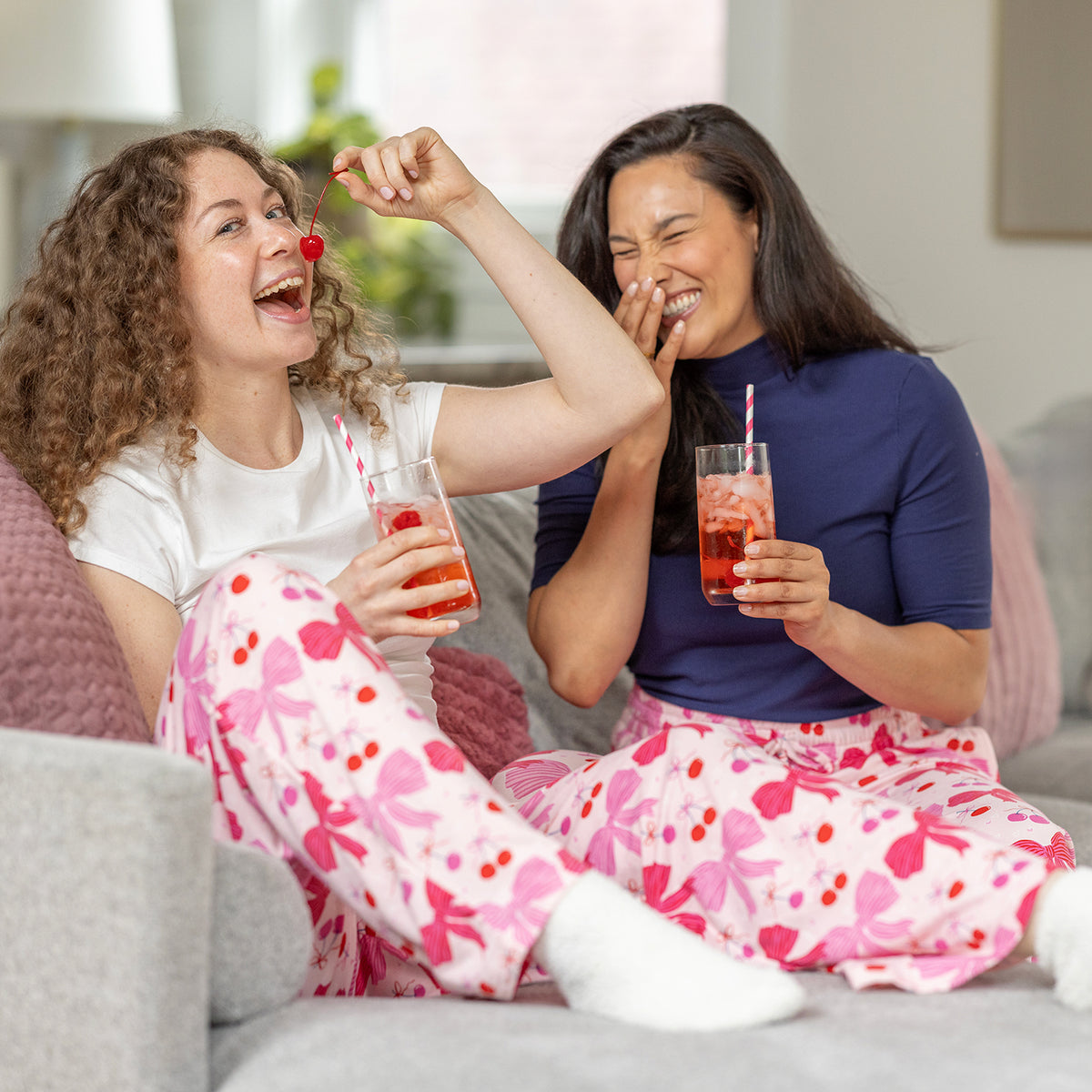 Two women in pajama pants sitting on a couch, enjoying drinks and a cherry.