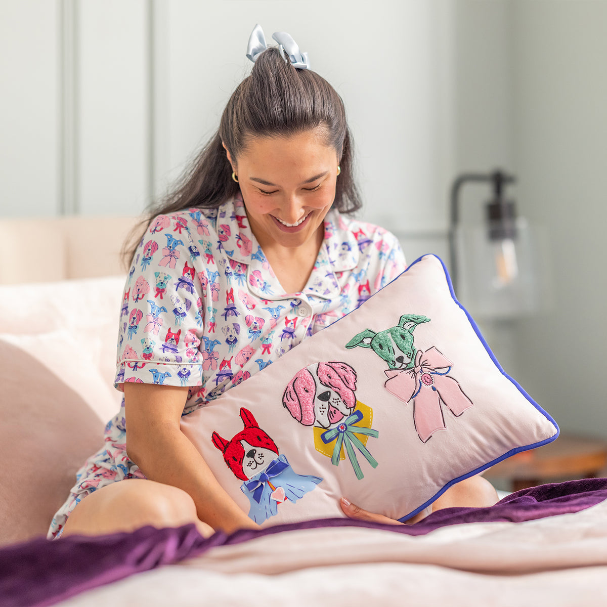Woman in pajamas holding a decorative pillow with cartoon designs in a bedroom setting.