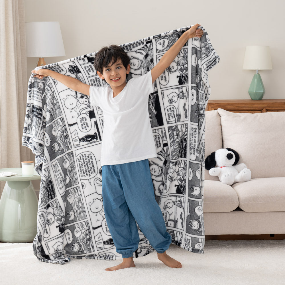 A child standing in a room, holding a black-and-white throw blanket with Peanuts comic strip illustrations.