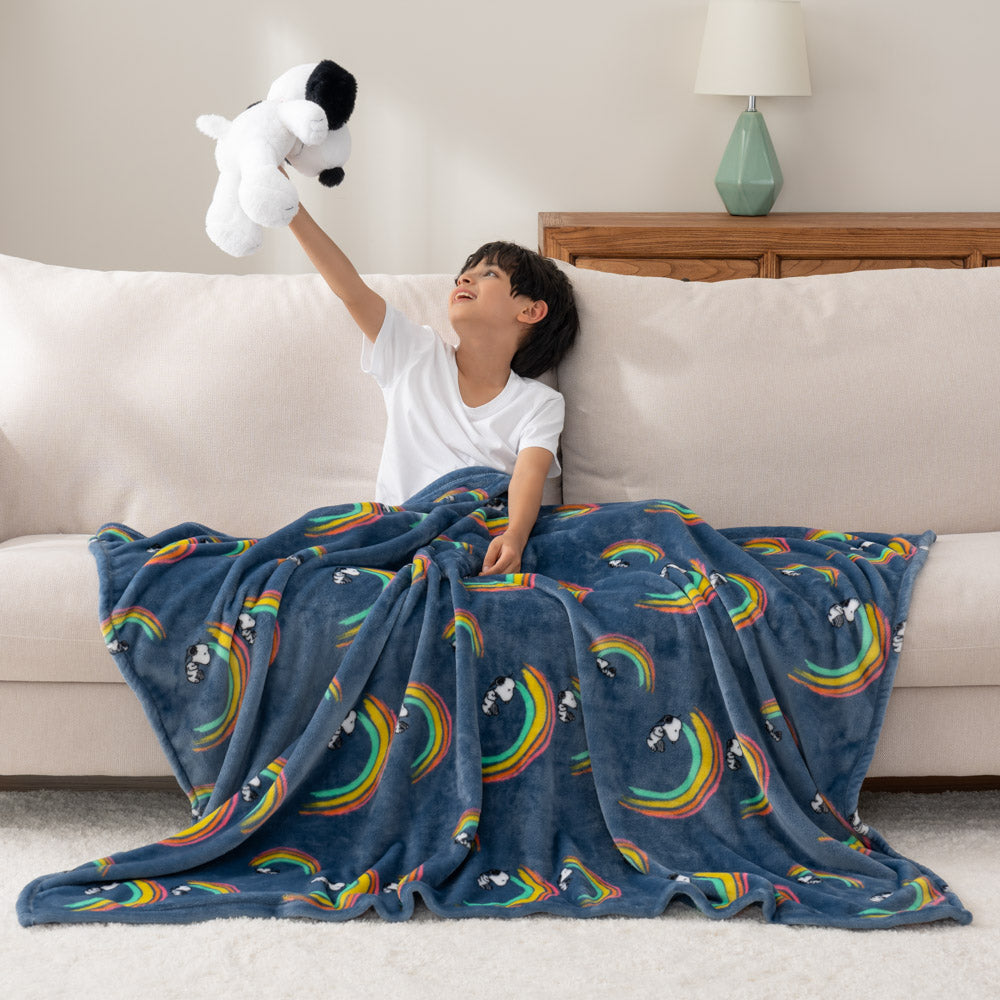 A child sitting on a sofa, wrapped in a blue throw blanket with snoopy under a rainbow as he holds up a snoopy plush toy.