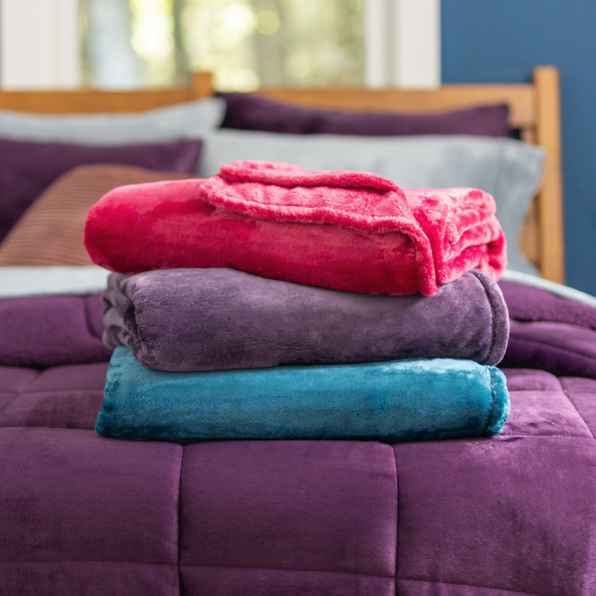 Image of a stack of VelvetLoft throws in hot pink, purple, and electric blue, on an eggplant purple bed.