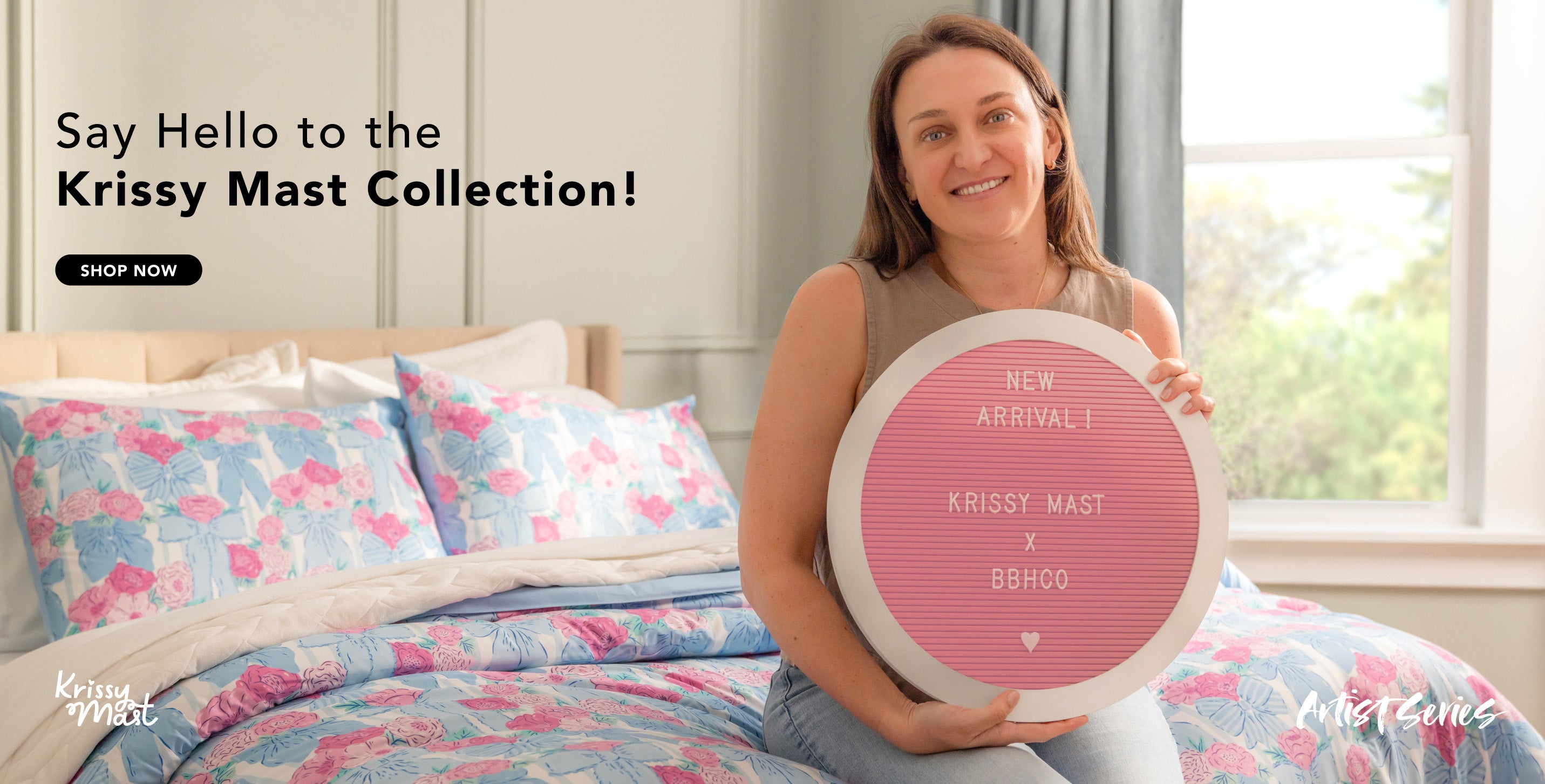 Say Hello to the Krissy Mast Collection! Shop Now! Image of artist, Krissy Mast, sitting on the foot of a bed that is styled with her product collection.