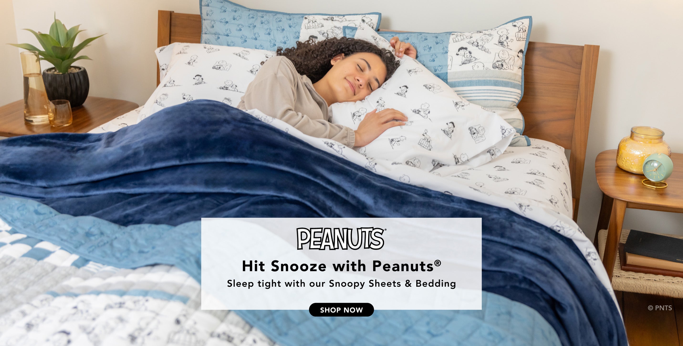Title: Hit Snooze with Peanuts®
Sub-Title: Sleep tight with our Snoopy Bedding
Image: Woman tucked into bed styled with black and white sheets, a blue blanket, and a quilt.