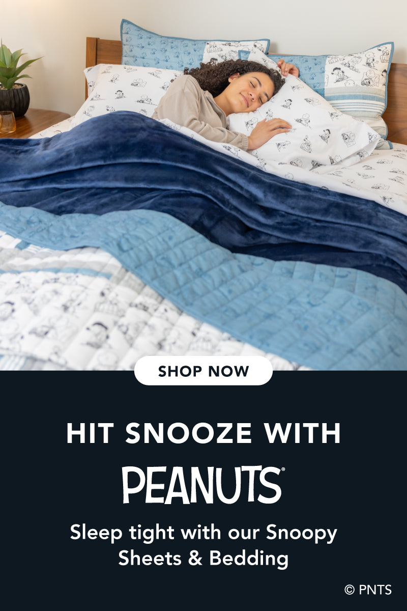 Title: Hit Snooze with Peanuts®
Sub-Title: Sleep tight with our Snoopy Bedding
Image: Woman tucked into bed styled with black and white sheets, a blue blanket, and a quilt.