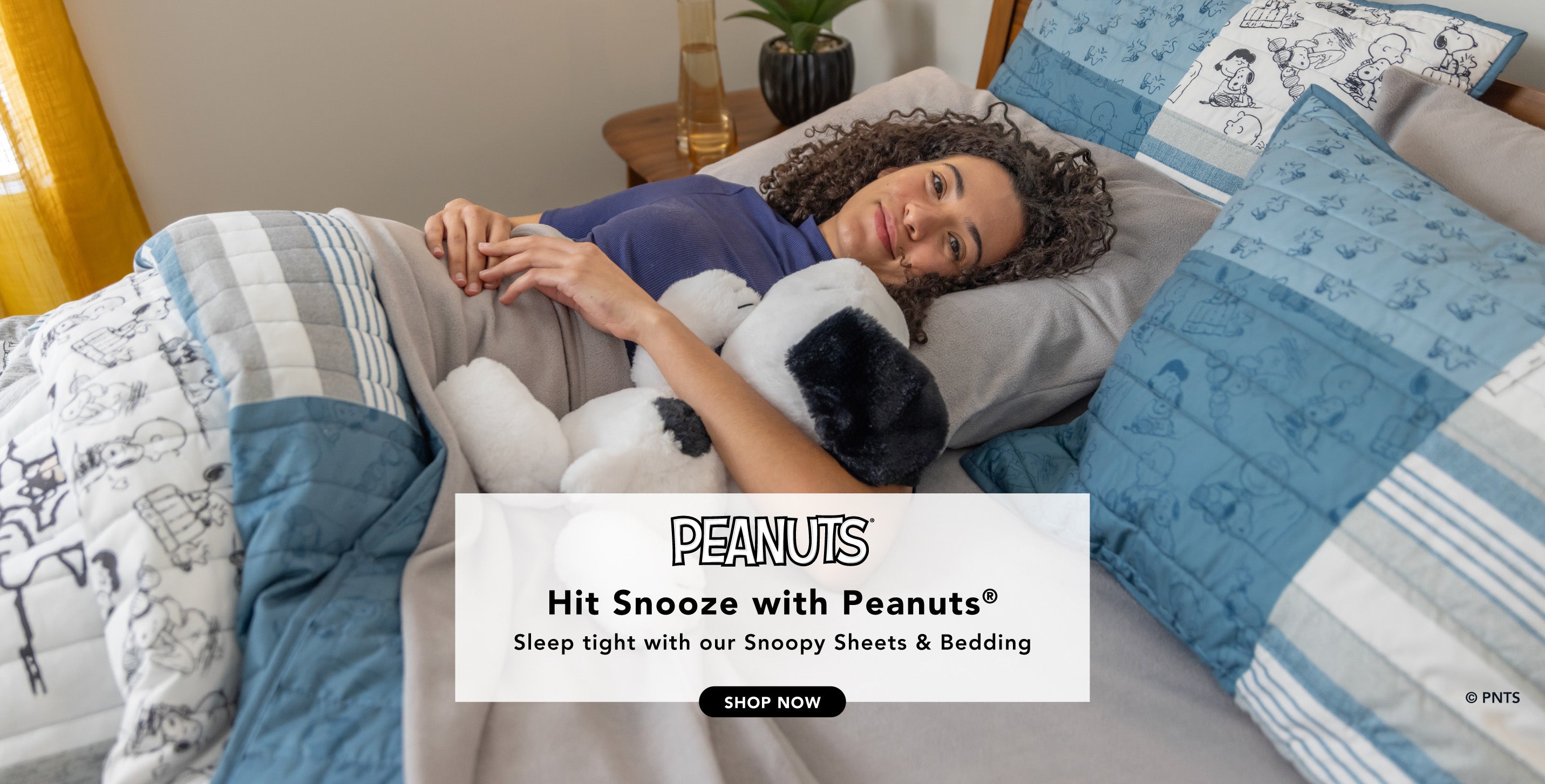 Title: Hit Snooze with Peanuts®
Sub-Title: Sleep tight with our Snoopy Bedding
Image: Woman tucked into bed styled with grey microfleece sheets and a Peanuts® quilt.