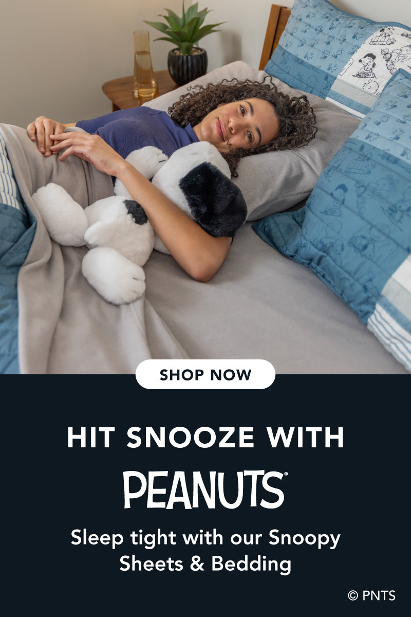 Title: Hit Snooze with Peanuts®
Sub-Title: Sleep tight with our Snoopy Bedding
Image: Woman tucked into bed styled with grey microfleece sheets and a Peanuts® quilt.
