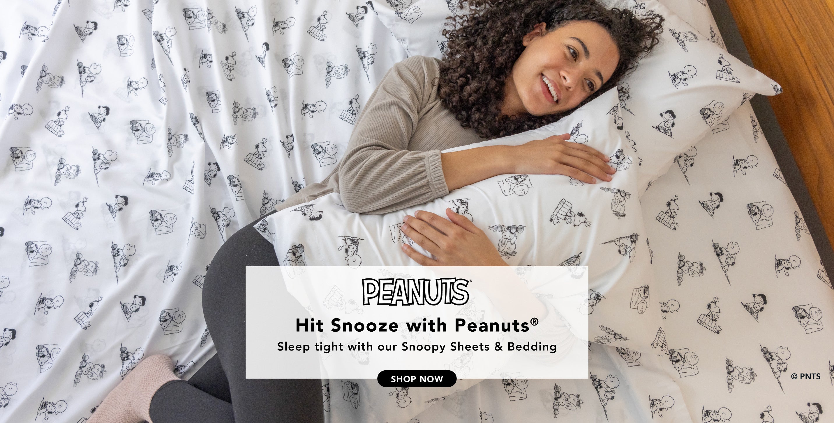 Title: Hit Snooze with Peanuts®
Sub-Title: Sleep tight with our Snoopy Bedding
Image: Woman tucked into bed styled with black and white sheets, a blue blanket, and a quilt.