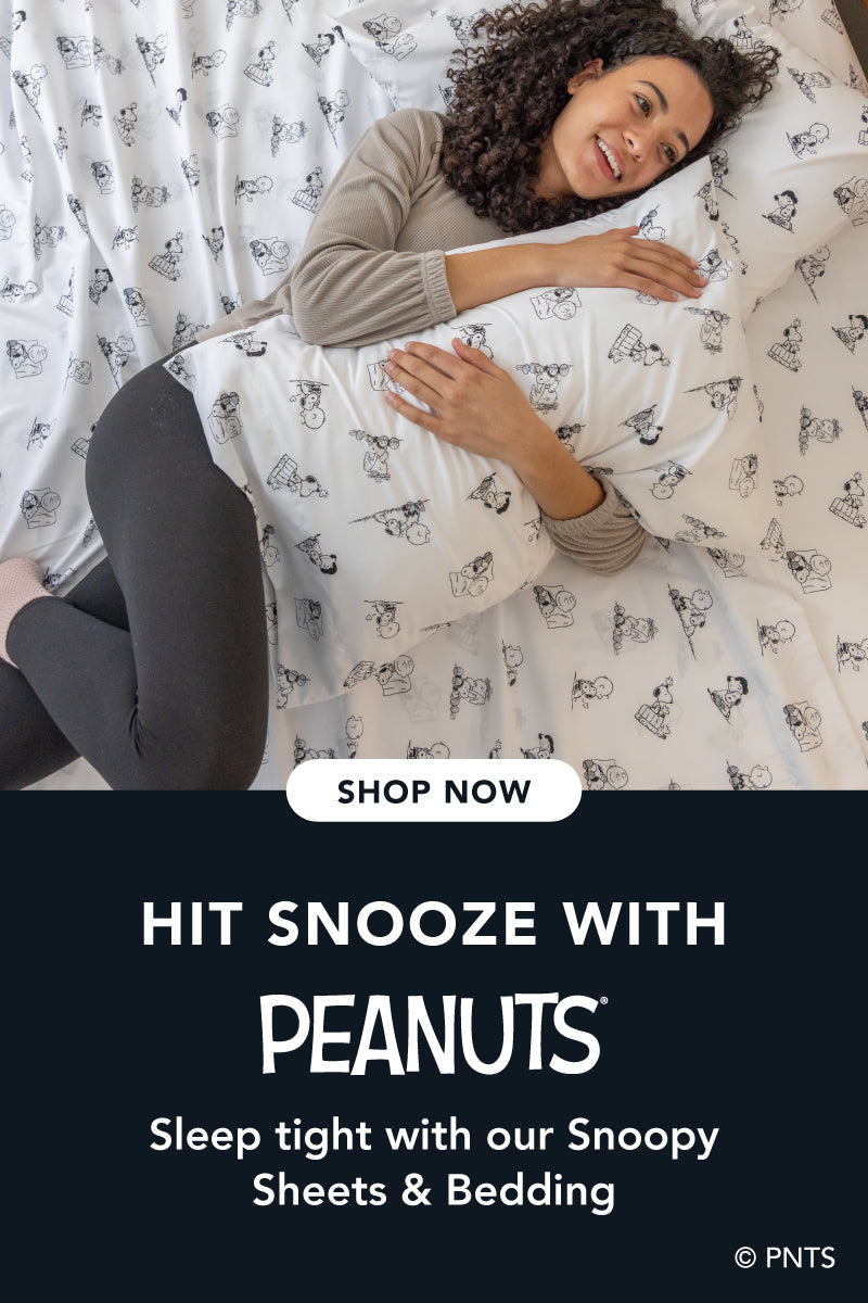 Title: Hit Snooze with Peanuts®
Sub-Title: Sleep tight with our Snoopy Bedding
Image: Woman tucked into bed styled with black and white sheets, a blue blanket, and a quilt.