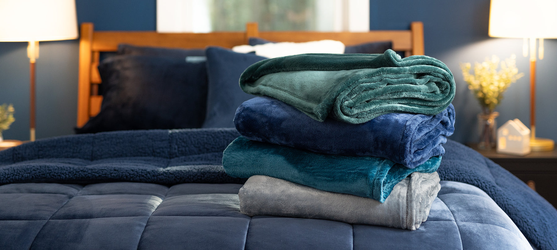 A stack of VelvetLoft throws sitting on a bed in a bedroom with our navy VelvetLoft Sherpa Comfortoer set and navy VelvetLoft sheets. 