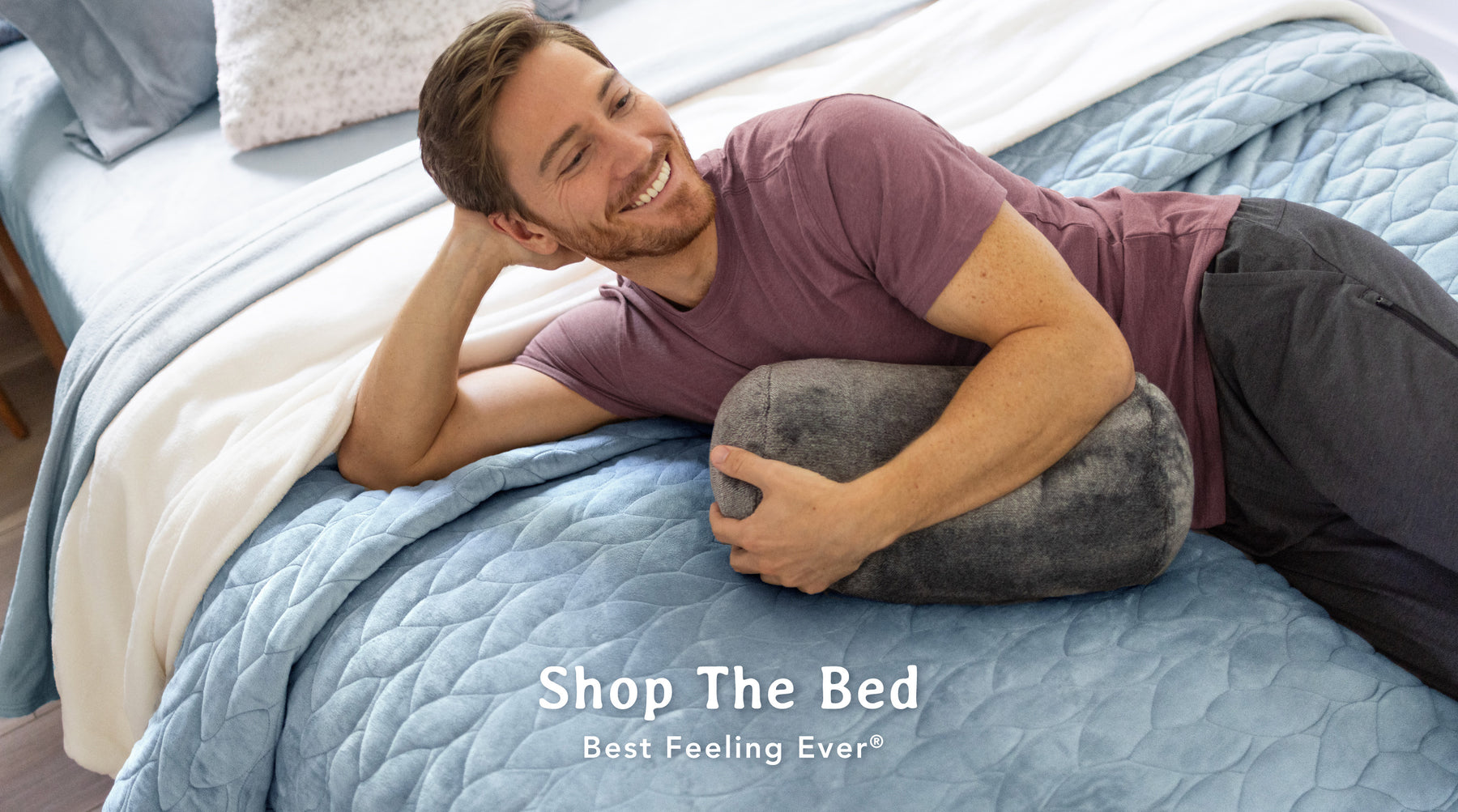 Image of a man casually laying atop a bed that is styled with a light blue VelvetLoft quilt, a white VelvetLoft blanket, and light blue VelvetLoft sheets. He is cuddling a pillow and smiling. 