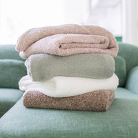 Stack of folded Retro comfort plush throws on top of a green couch. The throw colors in the pile from top to bottom are: Hushed Auburn, Farmhouse Sage, White, Sumatra Blend