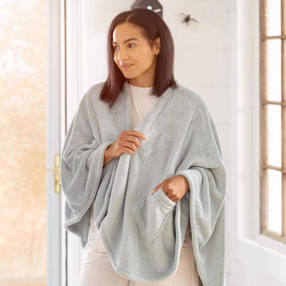 A woman standing in a bright room, wearing a light blue PrimaLush Cape Wrap with her hand tucked into the pocket.