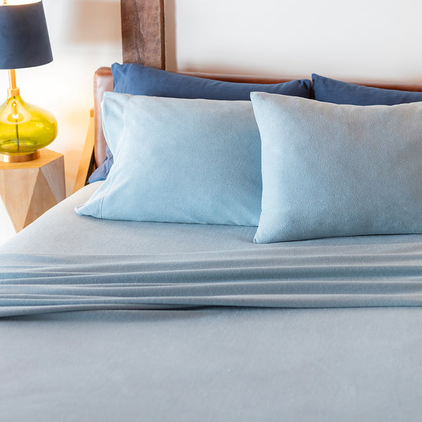 A neatly made bed with a light blue sheet and matching blue pillows, next to a yellow lamp.