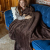 A woman sitting on a blue sofa, wrapped in a brown Extra-Fluffy Throw and smiling.