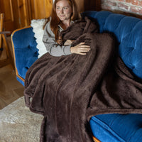 A woman sitting on a blue sofa, wrapped in a brown Extra-Fluffy Throw and smiling.