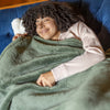 A woman lying on a blue sofa, smiling while wrapped in a sage green Extra-Fluffy Throw.