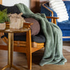 A sage green and brown Extra-Fluffy Throw draped over a wooden chair in a cozy living room, next to a small table with decorative items.