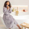 Woman in a lilac robe standing by a bathtub in a bathroom setting.