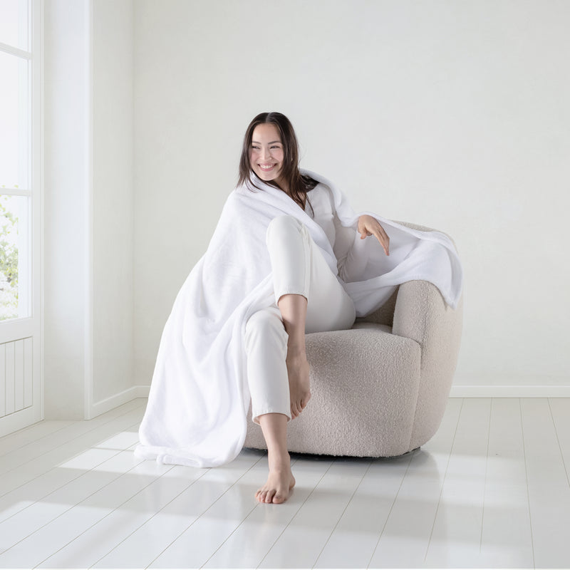 A woman sitting on a boucle chair with white velvety plush throw draped over her shoulders as she smiles.