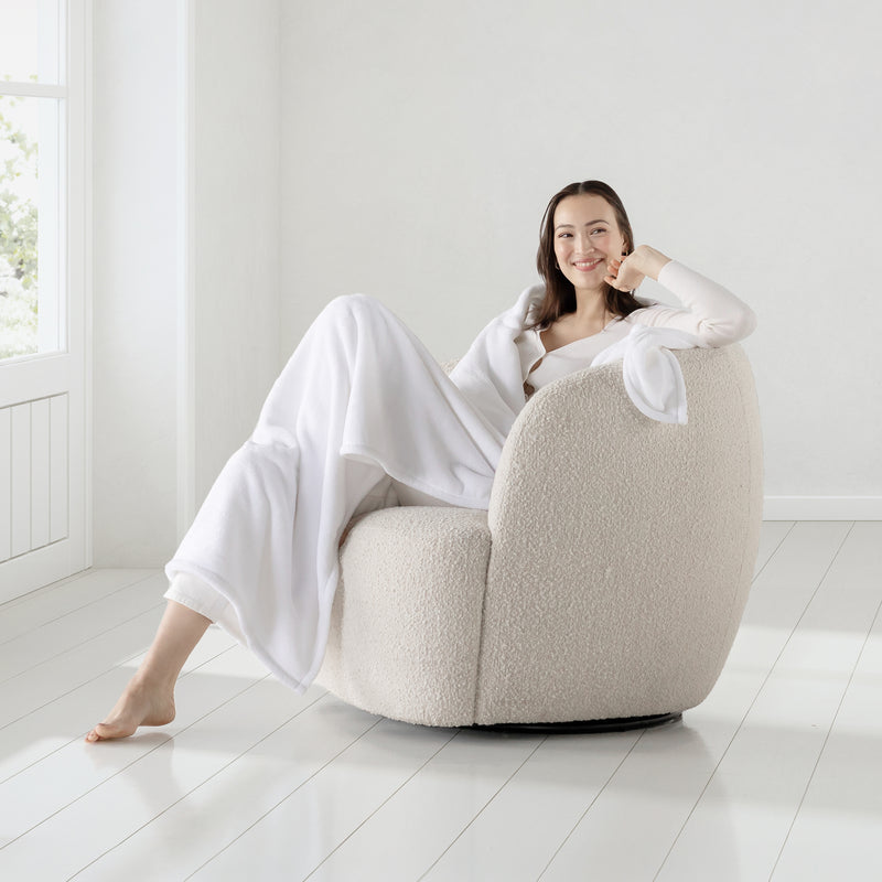 A woman sitting on a boucle chair with white velvety plush throw draped over her shoulders as she smiles.