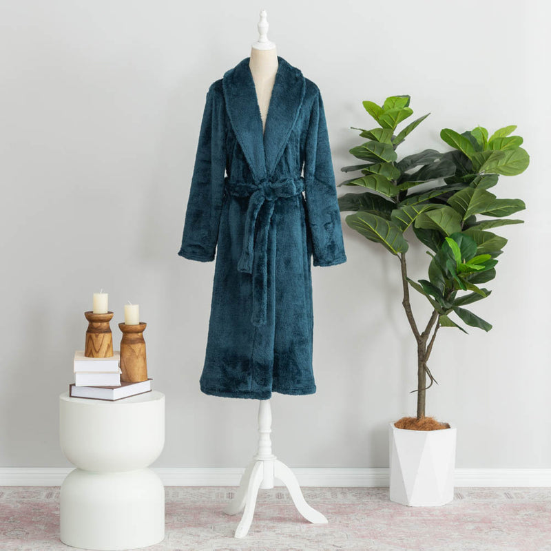 Juniper Stone-colored plush Extra Fluffy robe displayed on a white mannequin stand. The robe is tied at the waist, styled in a minimalist setting with a potted green plant on one side and a white side table holding books, wooden candle holders, and candles on the other.

