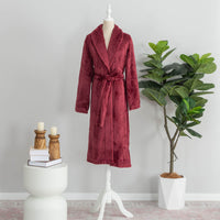 Windsor Wine-colored plush Extra Fluffy robe displayed on a white mannequin stand. The robe is tied at the waist, styled in a minimalist setting with a potted green plant on one side and a white side table holding books, wooden candle holders, and candles on the other.
