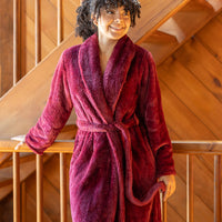 Woman standing on a wooden staircase landing, wearing a plush extra-fluffy robe in Windsor Wine color. Robe is  tied at the waist. She smiles softly while looking off to the side. The natural wood-paneled setting adds warmth to the scene.