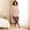 A woman standing in a bright bedroom, wearing a blush pink PrimaLush Cape Wrap styled with gray leggings.