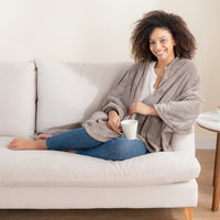 A woman reclining on a sofa with a mug, wrapped in a taupe PrimaLush Cape Wrap.