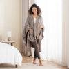 A taupe PrimaLush Cape Wrap worn by a woman standing in a bright, modern room.