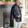 A man standing by a fireplace, smiling while wearing a black PrimaLush Cape Wrap.