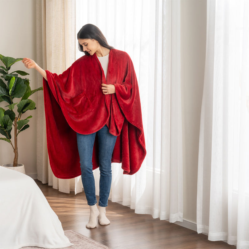 A woman standing in a bright room, wearing a red PrimaLush Cape Wrap styled over casual clothing.