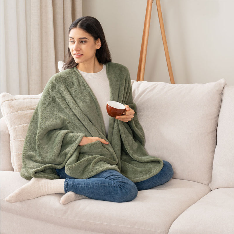 A woman sitting on a sofa while holding a mug, wearing an olive green PrimaLush Cape Wrap, smiling comfortably.